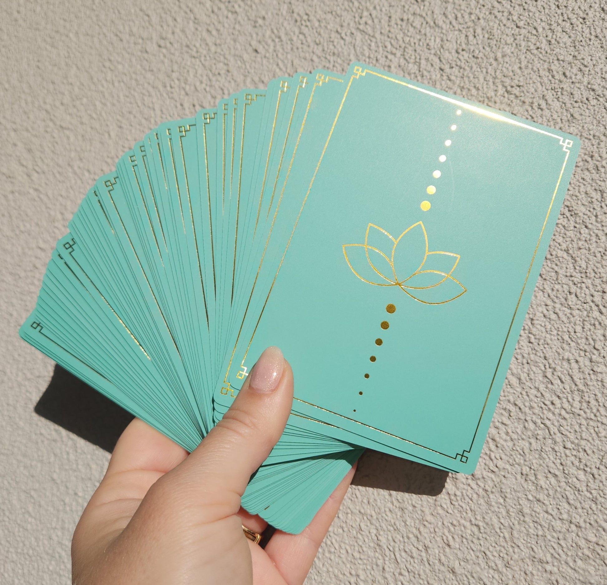 Hand holding a deck of teal cards with a gold lotus design on a neutral background